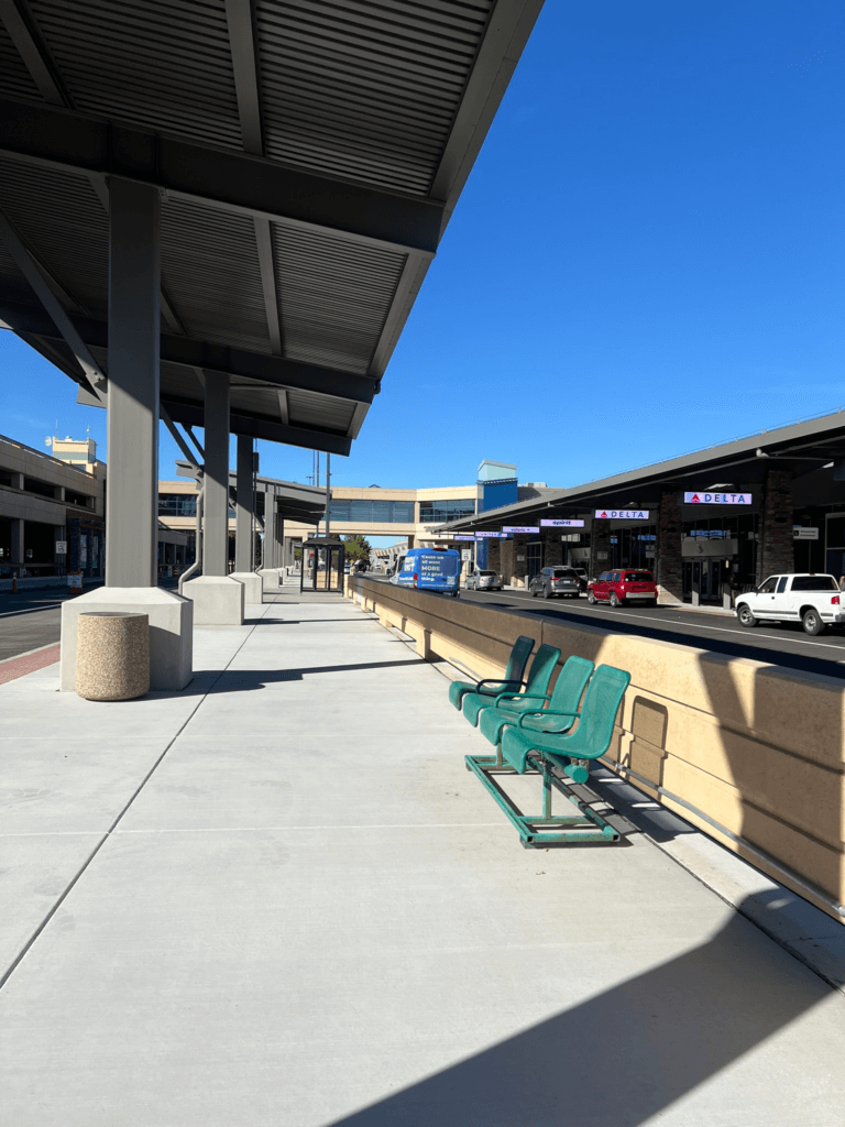 View of passenger drop off curb with shade structure, sidewalk and seating.